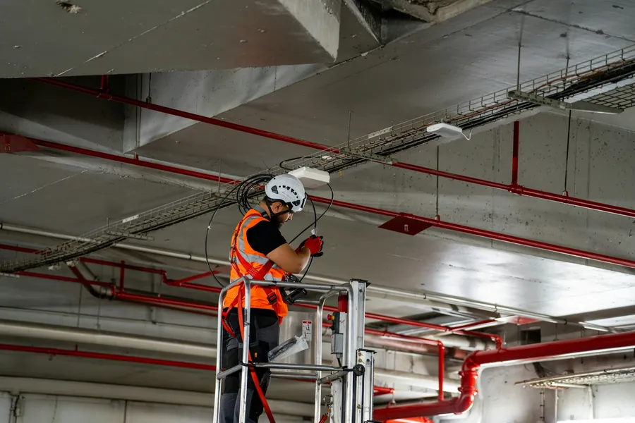 Technician in a hard hat and safety vest working on overhead cable trays from a lift in a commercial ceiling